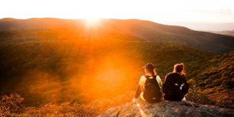 Two people sitting on a mountain with the sun setting in front.
